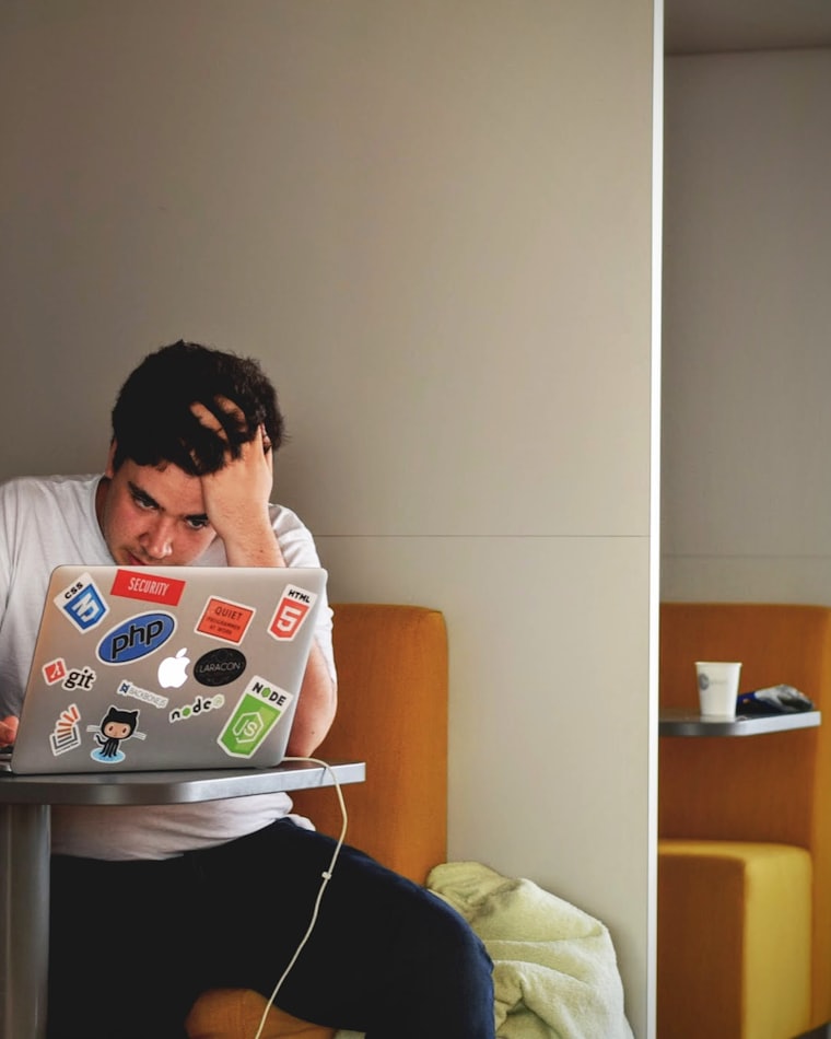 A man at a desk holding his forehead in front of a laptop