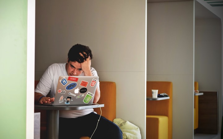 A man at a desk holding his forehead in front of a laptop